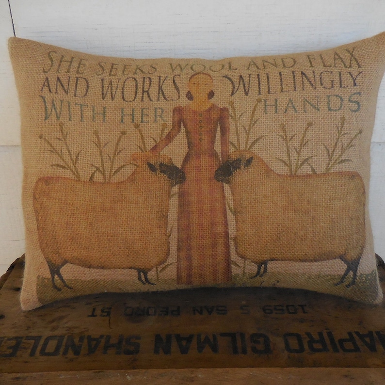 May include: A burlap pillow with a printed design of a woman in a long dress standing between two sheep. The text on the pillow reads "She seeks wool and flax and works willingly with her hands".