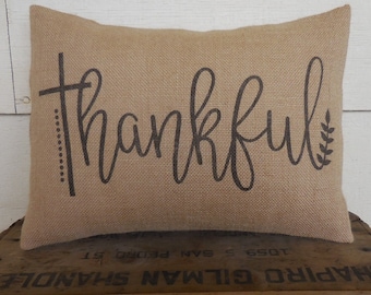 May include: A brown burlap pillow with the word "thankful" printed in black script. The pillow has a simple design with a small leaf sprig on the right side.