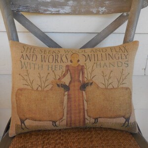 May include: A burlap pillow with a printed design of a woman standing between two sheep. The text on the pillow reads "She seeks wool and flax and works willingly with her hands."