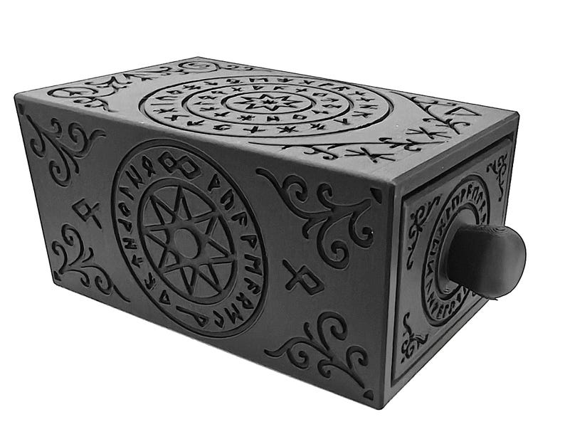May include: A black rectangular box with intricate, embossed designs. The box features circular and linear patterns with symbols and a central star design. A round knob is on the front. The box has a mysterious, ancient aesthetic.