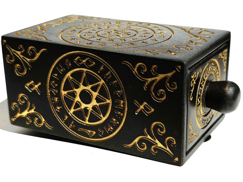 May include: Black wooden box with gold-colored etched designs. The box features circular patterns, starbursts, and ornate scrollwork. A dark wooden knob is on the side. The box has a mysterious, antique aesthetic.