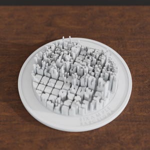 May include: A white circular 3D model of the Eixample district in Barcelona, featuring a detailed relief map of city blocks and buildings. The base is a smooth white disc with the text EIXAMPLE BARCELONA embossed on the lower right edge.