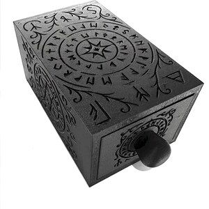 May include: A dark gray rectangular box with intricate carvings of runes and floral patterns. The box features a central star design and a circular pattern on the front. A black knob is visible on the front.