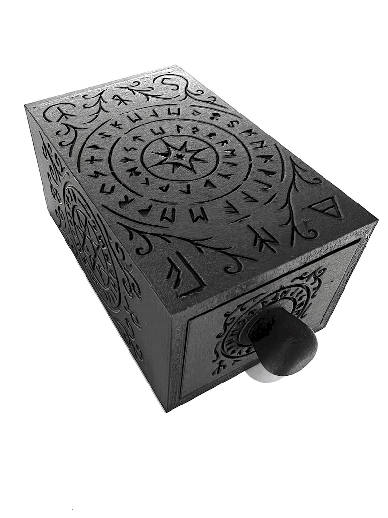 May include: A dark gray rectangular box with intricate carvings of runes and floral patterns. The box features a central star design and a circular pattern on the front. A black knob is visible on the front.