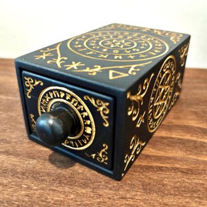 May include: A black wooden box with a drawer, adorned with gold-colored circular and floral designs. The box features a knob on the drawer and intricate patterns on the sides and top, creating a decorative and detailed appearance.
