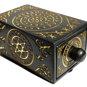 May include: A black rectangular box with intricate gold-colored carvings. The design includes circular patterns, stars, and swirling lines. A dark knob is on the front. The box is likely a decorative item or a container.