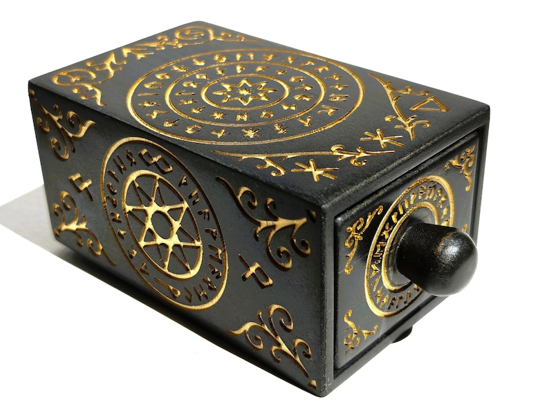 May include: A black rectangular box with intricate gold-colored carvings. The design includes circular patterns, stars, and swirling lines. A dark knob is on the front. The box is likely a decorative item or a container.