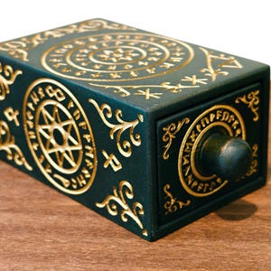 May include: A dark teal rectangular box with a drawer, adorned with gold-colored, intricate, circular and spiral designs. The box features a small, round knob and is set on a wooden surface.