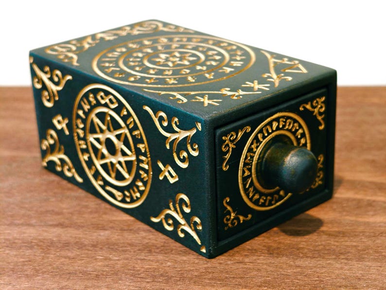 May include: A dark teal rectangular box with a drawer, adorned with gold-colored, intricate, circular and spiral designs. The box features a small, round knob and is set on a wooden surface.