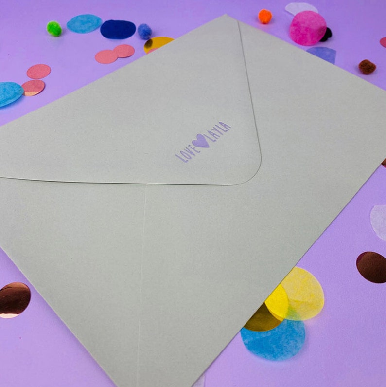 May include: A light green envelope with the text "LOVE LAYLA" and a heart symbol printed in purple. The envelope is surrounded by colorful confetti on a purple surface.