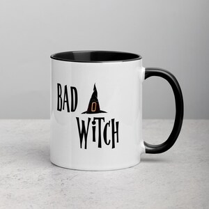 May include: White ceramic mug with a black interior and handle. The mug features the words "BAD WITCH" in black text, with a witch hat graphic. A Halloween-themed mug.