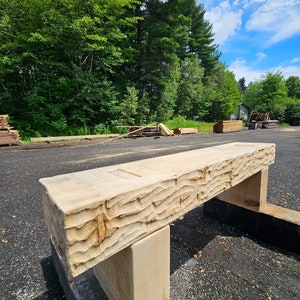 Könnte beinhalten: Eine rustikale Holzbank aus einem einzigen Stück Holz mit einer natürlichen, grob behauenen Oberfläche. Die Bank hat zwei einfache Holzstützen und steht auf einer Kiesfläche.