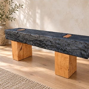 May include: A bench featuring a long, rectangular seat with a dark, textured, charcoal-colored finish and two light-toned, natural wood block legs. The seat includes two small, rectangular wooden inlays on the top surface, providing a contrast.