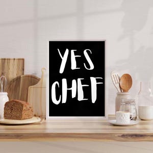 May include: Black and white kitchen print with the text "YES CHEF" in white lettering on a black background. The print is framed in a white frame and is sitting on a wooden countertop.