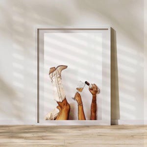 May include: A framed art print featuring a watercolor illustration. The artwork depicts a pair of white cowboy boots, a person holding a cocktail glass, and another person pouring a drink from a bottle. The color palette includes shades of brown, white, and gold.
