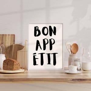 May include: A white framed print with black text that reads "Bon Appétit" on a wooden countertop with kitchen utensils and a loaf of bread.