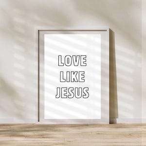 May include: A framed white print with the words "LOVE LIKE JESUS" in bold, black letters. The frame is a light gray color. The print is leaning against a white wall with a wooden floor.