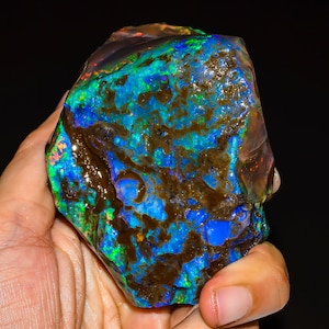 May include: A raw boulder opal specimen featuring a vibrant play of color with intense electric blue and green flashes interspersed with dark brown ironstone matrix veins. The surface displays a polished, irregular texture with scattered red highlights.