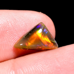 May include: A triangular-shaped loose gemstone featuring a dark base with vibrant flashes of orange, yellow, and blue iridescence. The stone has a smooth, polished surface that reflects light, showcasing a play of colour across its faceted edges.