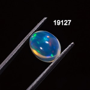Puede incluir: Una vibrante piedra preciosa de ópalo de forma ovalada, sujeta con pinzas sobre un fondo negro. El ópalo muestra un cautivador juego de colores, con destellos de azul, verde y amarillo. El número "19127" es visible en texto blanco.