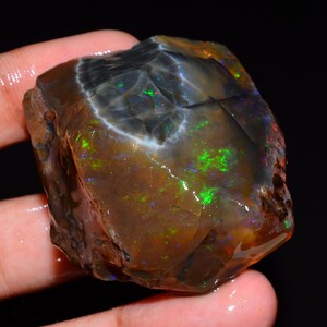 May include: A raw, unpolished boulder opal gemstone featuring a dark, translucent brown base with vibrant flashes of neon green, blue, and red play-of-color throughout its irregular, textured surface against a solid black background.