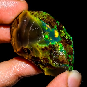 May include: A raw, unpolished opal gemstone featuring a dark, translucent base with vibrant flashes of neon green and golden yellow play-of-color. The stone has an irregular, organic shape with a smooth, glassy section and a rough, textured surface.