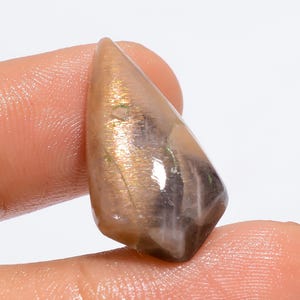 May include: A polished, teardrop-shaped gemstone with a mix of colors. The stone has a translucent appearance with shades of brown, gold, and hints of green. The gem is held between two fingers, showcasing its unique shape and natural beauty.