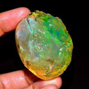May include: A raw, unpolished opal gemstone displaying a vibrant play-of-color with dominant flashes of neon green, yellow, and orange against a translucent, milky white base. The stone has an irregular, organic shape with a textured surface.
