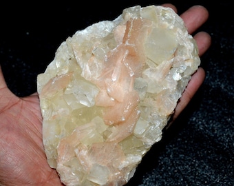 Op de afbeelding: Een natuurlijke kristalformatie met een mix van lichtgele en perzikkleurige kristallijne structuren. Het mineraalmonster wordt in een hand gehouden, waarbij de onregelmatige vorm en het getextureerde oppervlak worden getoond. Het kristal lijkt een geode of een vergelijkbaar type gesteente te zijn.