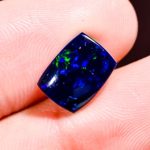 Peut inclure: Une pierre précieuse opale noire rectangulaire à la surface polie, présentant des éclats vibrants de bleu électrique et de vert vif dans la base bleu foncé de la pierre, avec un dessus lisse et légèrement bombé.