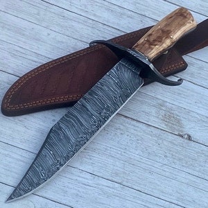 Pu&ograve; includere: Un grande coltello a lama fissa con un fodero in pelle marrone. La lama &egrave; in acciaio a motivi e l'impugnatura &egrave; in legno.