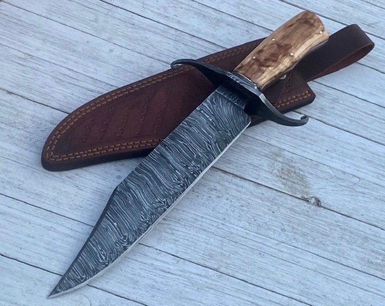 Pu&ograve; includere: Un grande coltello a lama fissa con un fodero in pelle marrone. La lama &egrave; in acciaio a motivi e l'impugnatura &egrave; in legno.