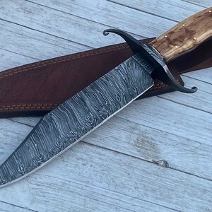 Pu&ograve; includere: Un coltello Bowie in acciaio Damasco con un fodero in pelle marrone e un manico in legno. Il coltello ha un motivo ondulato distintivo sulla lama.
