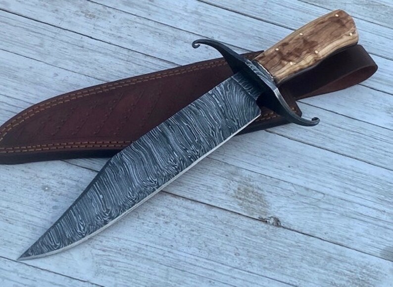 Pu&ograve; includere: Un coltello Bowie in acciaio Damasco con un fodero in pelle marrone e un manico in legno. Il coltello ha un motivo ondulato distintivo sulla lama.