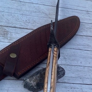 Pu&ograve; includere: Un coltello lungo color argento con un manico in legno. Il manico &egrave; di colore marrone chiaro e una striscia marrone scuro corre lungo il centro. Il coltello &egrave; appoggiato su un fodero in pelle marrone.