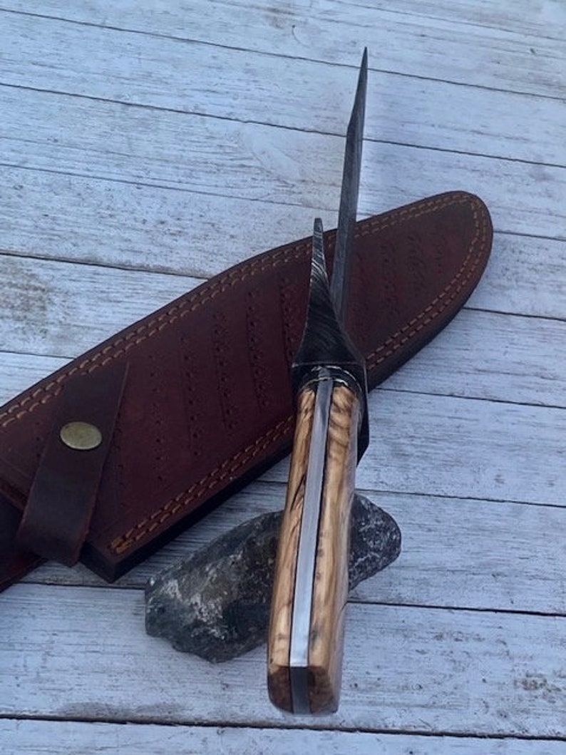 Pu&ograve; includere: Un coltello lungo color argento con un manico in legno. Il manico &egrave; di colore marrone chiaro e una striscia marrone scuro corre lungo il centro. Il coltello &egrave; appoggiato su un fodero in pelle marrone.