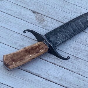 Pu&ograve; includere: Un coltello artigianale con manico in legno e lama in acciaio damascato. La lama presenta un caratteristico motivo ondulato e il manico &egrave; fissato con rivetti in ottone.