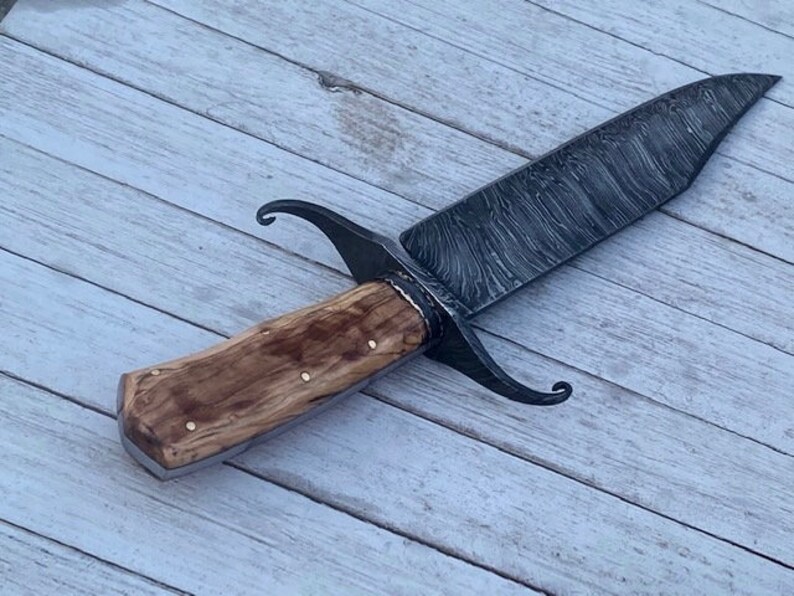 Pu&ograve; includere: Un coltello artigianale con manico in legno e lama in acciaio damascato. La lama presenta un caratteristico motivo ondulato e il manico &egrave; fissato con rivetti in ottone.