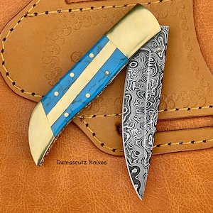 May include: A blue and brass pocket knife with a damascus steel blade. The knife is closed and resting in a leather sheath.