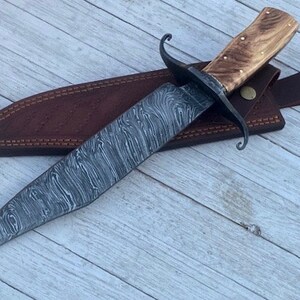 Pu&ograve; includere: Un coltello da caccia artigianale con una lama in acciaio damascato e un manico in legno. Il coltello &egrave; in un fodero di cuoio marrone con una cinghia.