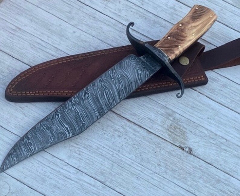Pu&ograve; includere: Un coltello da caccia artigianale con una lama in acciaio damascato e un manico in legno. Il coltello &egrave; in un fodero di cuoio marrone con una cinghia.