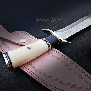 May include: A hunting knife with a white bone handle, black and red accents, and a silver blade with a decorative pattern. The knife is in a leather sheath with decorative stitching.