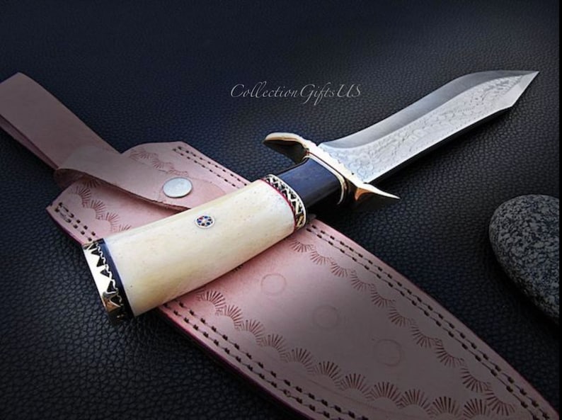 May include: A hunting knife with a white bone handle, black and red accents, and a silver blade with a decorative pattern. The knife is in a leather sheath with decorative stitching.