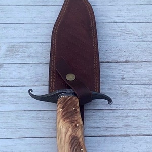 Pu&ograve; includere: Un fodero in pelle marrone con un coltello a lama curva con manico in legno marrone. Il coltello ha un accento in ottone sul manico.