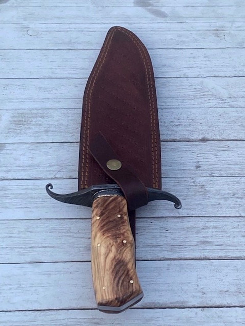 Pu&ograve; includere: Un fodero in pelle marrone con un coltello a lama curva con manico in legno marrone. Il coltello ha un accento in ottone sul manico.