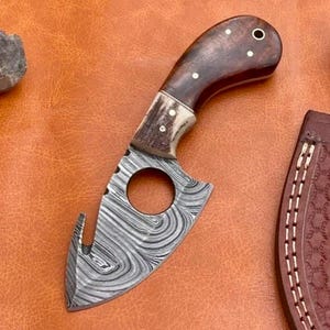 May include: A small, hand-forged knife with a Damascus steel blade and a brown wood handle. The blade has a hook on the end and a round hole in the center. The knife is sitting on a brown leather surface.