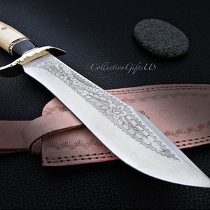 May include: A large, silver knife with a decorative etched design on the blade. The knife has a bone handle with a black inlay and a brass guard. The knife is resting on a brown leather sheath with decorative stitching.