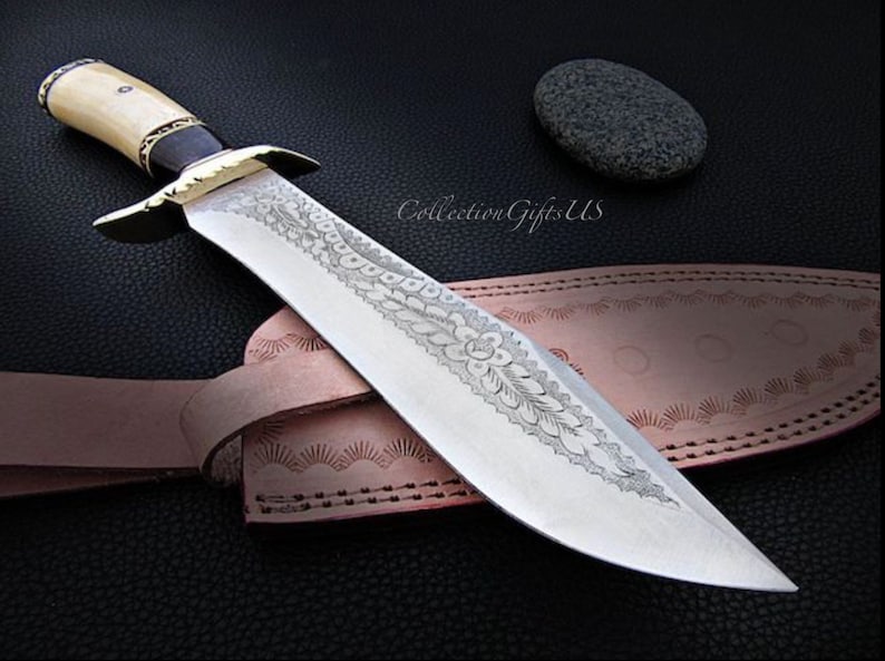 May include: A large, silver knife with a decorative etched design on the blade. The knife has a bone handle with a black inlay and a brass guard. The knife is resting on a brown leather sheath with decorative stitching.