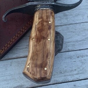 Pu&ograve; includere: Primo piano di un manico in legno con dettagli in ottone su un coltello da caccia. Il manico &egrave; in legno marrone chiaro con venature marroni scure. La lama non &egrave; visibile nell'immagine.