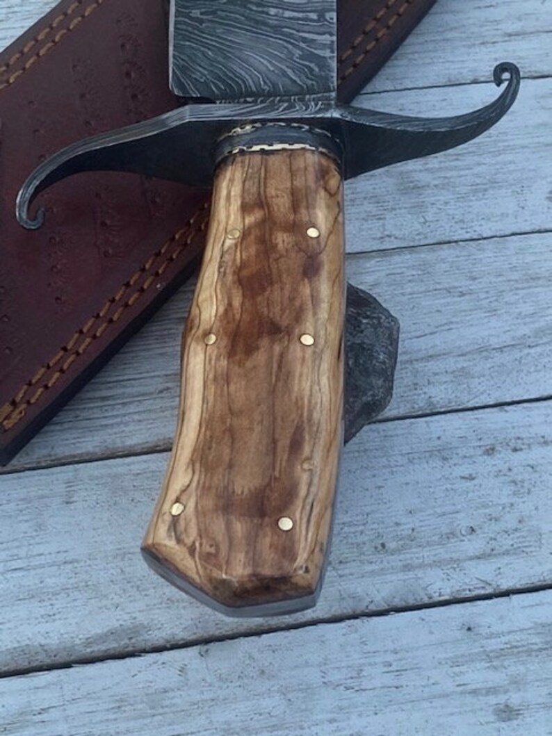Pu&ograve; includere: Primo piano di un manico in legno con dettagli in ottone su un coltello da caccia. Il manico &egrave; in legno marrone chiaro con venature marroni scure. La lama non &egrave; visibile nell'immagine.